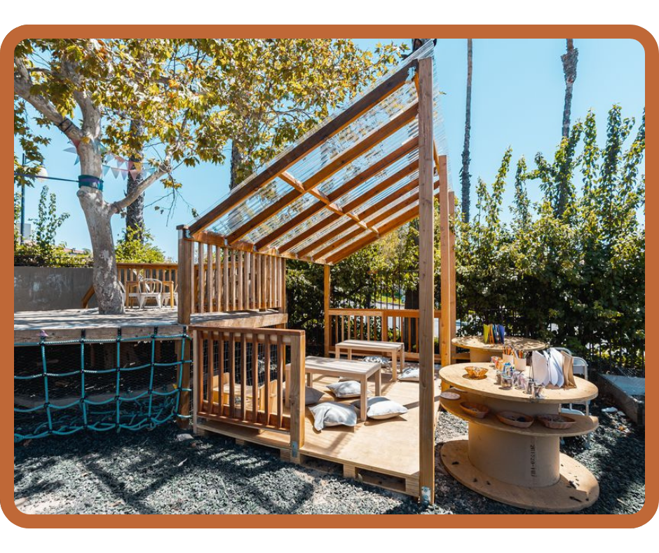 Outdoor preschool environment with wooden playhouse, seating cushions, and activity table inspired by Montessori and Reggio Emilia learning